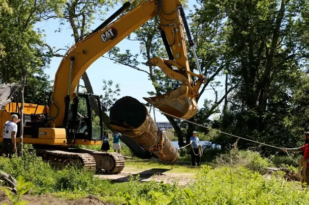 Large, yellow crane, picks up mishoon log for placement on the Historic Patuxet Homesite.