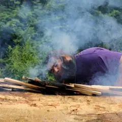 Museum educator blows onto embers of mishoon burn. Smoke rises dramatically.