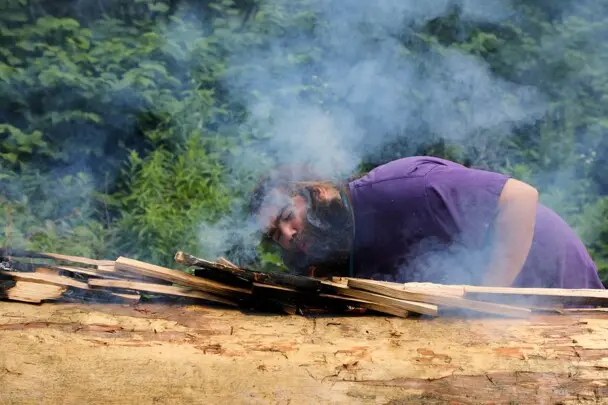 Museum educator blows onto embers of mishoon burn. Smoke rises dramatically.