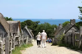 A couple walks down the main path of the 17th-Century English Village. Small, grey, clapboard houses flank the street and the ocean is before them in the distance.