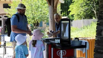 Cafe worker hands an ice cream cone to a young girl at an ice cream cart with her family.