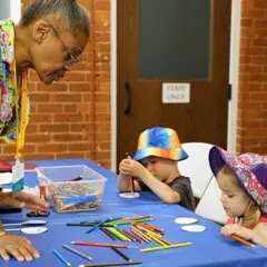 Young children sit at a craft table with a Museum volunteer guides them in crafts.
