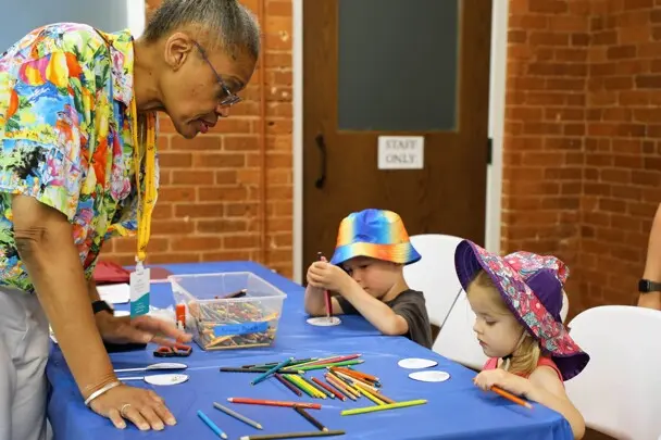 Young children sit at a craft table with a Museum volunteer guides them in crafts.