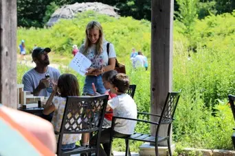 A family pauses to play giant Jenga, eat icea cream, and look at a scavenger hunt at an outdoor table. Tall green grass and a wetu are behind them in the distance.