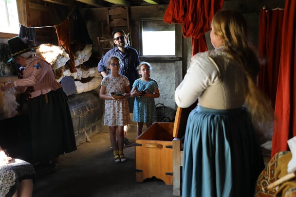Two young girls watch the Pilgrim Bride as she gets ready inside of a house.