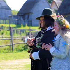 Pilgrim Bridge holds her groom's arm. Grey, clapboard houses with thatched roofs are behind them.