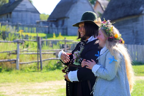 Pilgrim Bridge holds her groom's arm. Grey, clapboard houses with thatched roofs are behind them.