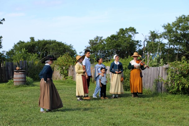 Guests mingle with Pilgrim women who hold musical instruments.