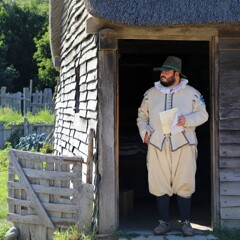 Pilgrim man stands in doorway of a grey, clapboard house.