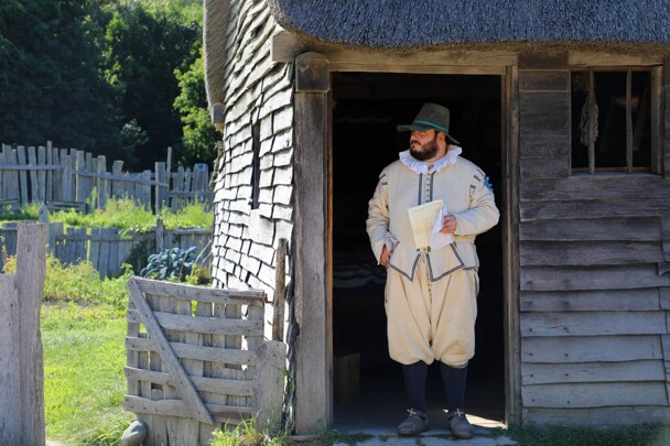 Pilgrim man stands in doorway of a grey, clapboard house.