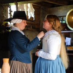 A Pilgirm woman helps the bride with her collar inside a house.