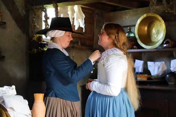 A Pilgirm woman helps the bride with her collar inside a house.