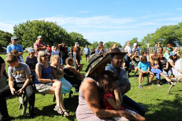 Museum guests gather as guests of a Pilgrim wedding on the Village Green. Most are seated, some stand.