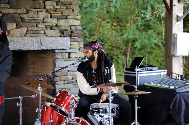 A member of The GroovaLottos plays the drums while singing into a microphone.