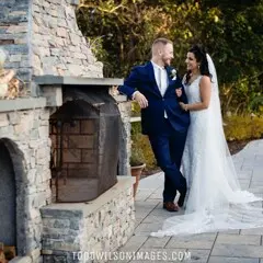 A bride and groom stand near a stone fireplace at an outdoor venue