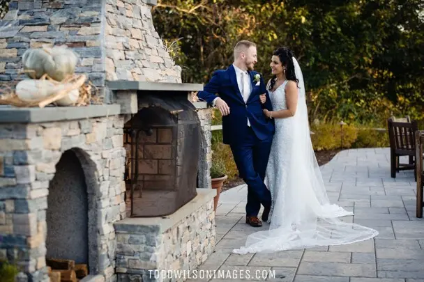 A bride and groom stand near a stone fireplace at an outdoor venue
