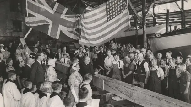 Crowd watches man hammer boat plank underneath the British and American flags