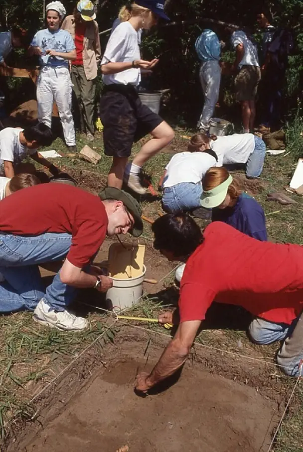 Several archeologists are in a field where some actively dig and others mill about. Multiple square shaped holes are in the ground.