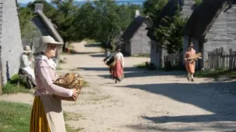 Pilgrim woman holds basket of corn on a road lined by grey shingle homes Additional Pilgrims perform daily chores in the background.