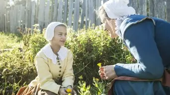 A pilgrim mother and daughter crouch in a garden while talking and picking flowers