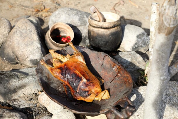 Roast chicken in a wooden bowl is set over stones. Cooking pots are in the background.