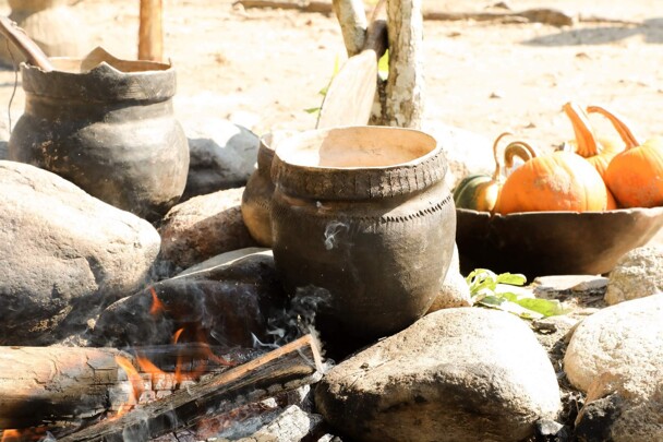 Wampanoag cooking pots set over an outdoor fire. A bowl with orange pumpkins is in the background.