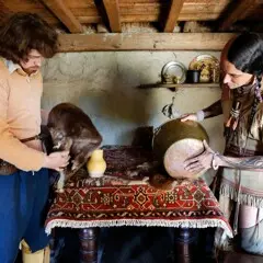 A pilgrim man and Wampanoag man inspect objects for trade in a home interior